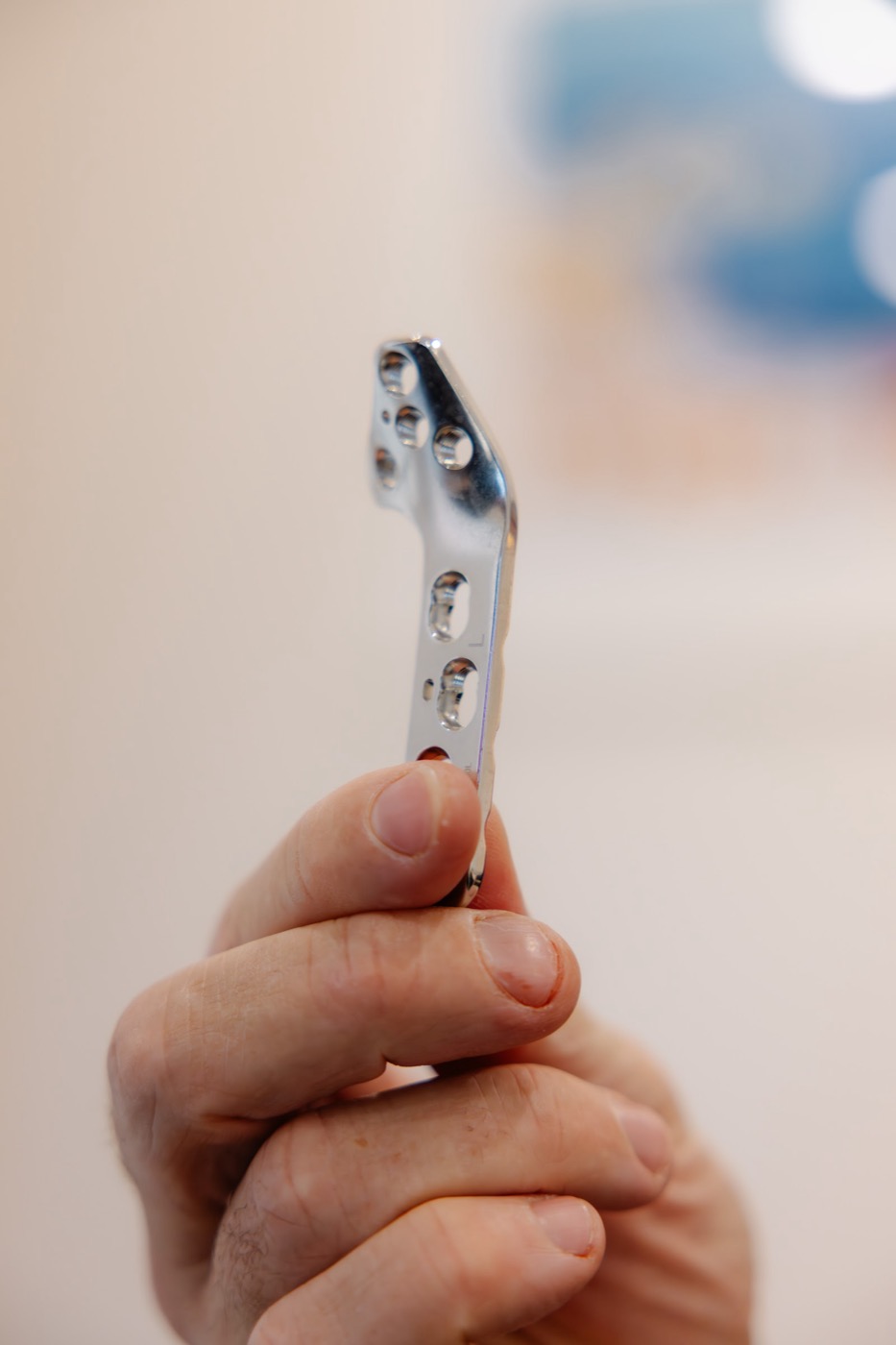 A hand holding up a polished stainless steel bone plate with countersunk screw holes